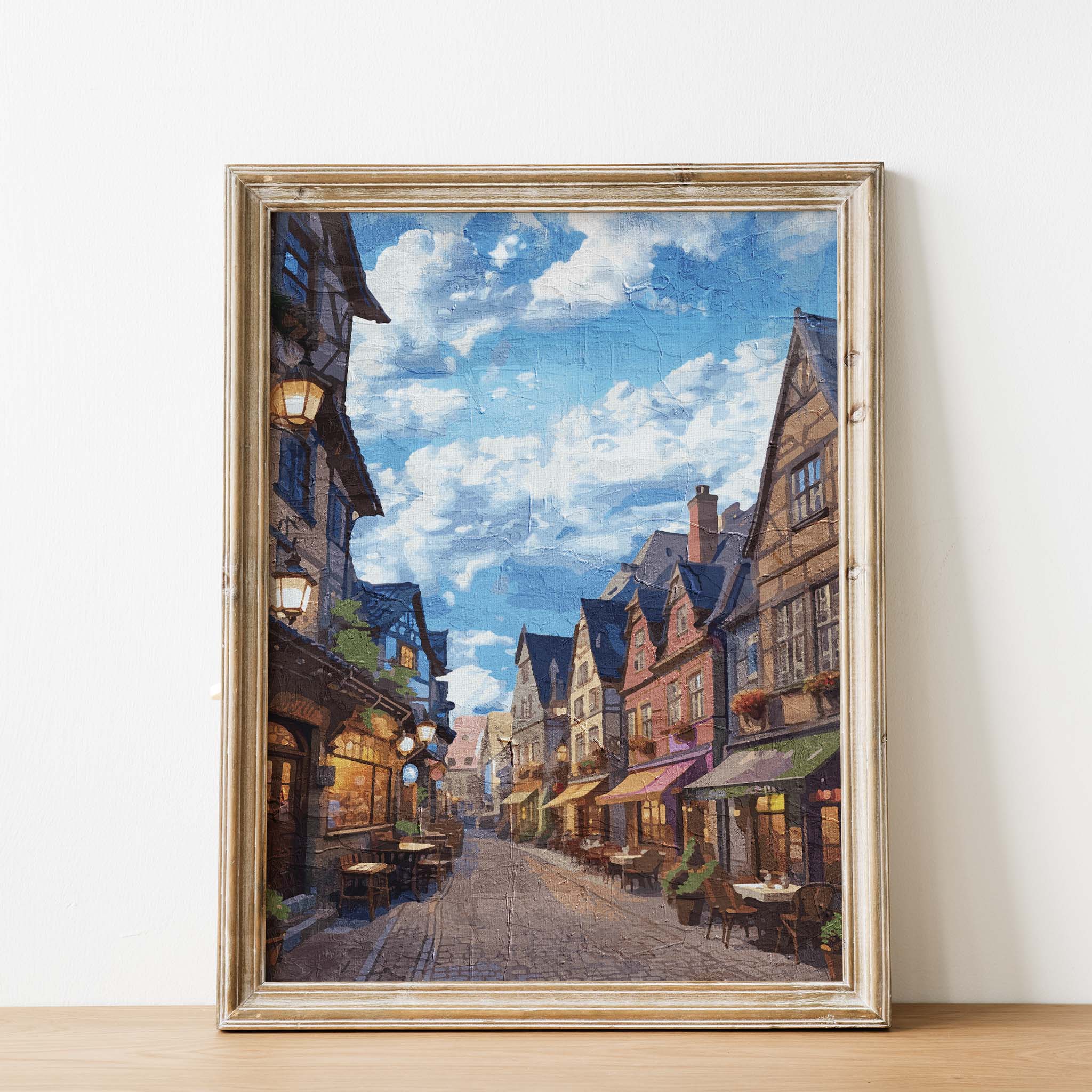 Cozy European village street wall art depicting a cobblestone alley lined with colorful historic buildings, quaint outdoor cafés, and warm lights under a vibrant blue sky with scattered clouds