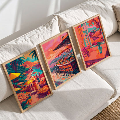 Three framed travel and lifestyle wall art prints on a wood shelf against sage green panelling — a tropical beach fruit market, a coastal bar with mountain views, and a warm interior window scene with flowers and a teal vase in vivid orange, teal, and coral