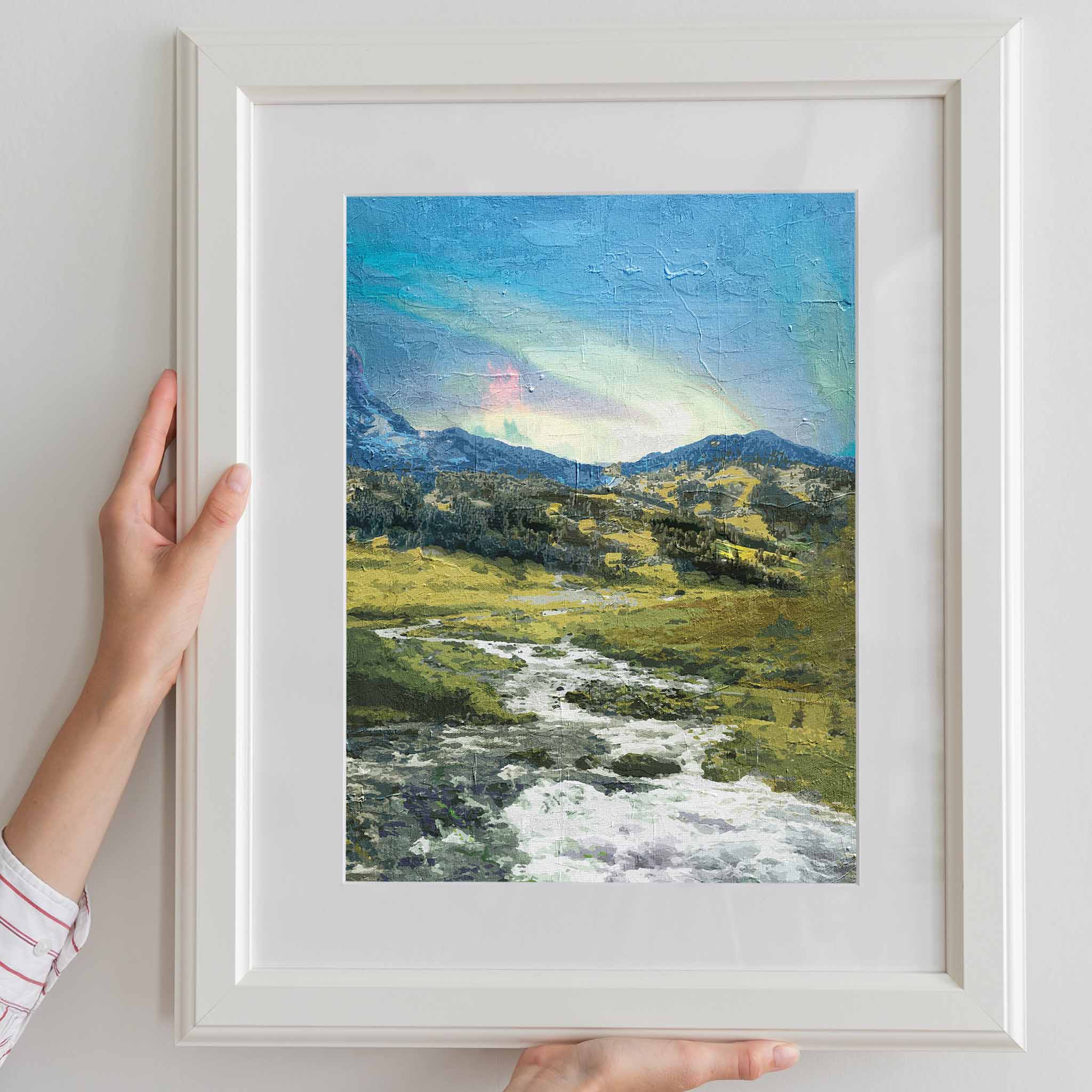 Rustic mountain valley wall art featuring a flowing stream winding through grassy hills and rocky terrain, with forested slopes and a soft, colorful sky above the distant peaks