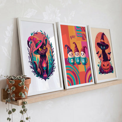 Halloween wall art prints on a wood shelf against sage green panelling - a witchy cat familiar print, a kawaii "Boo!" ghost trio in teacups, and a classic black cat on a jack-o-lantern with witch hat and bats in bold teal, orange, and magenta