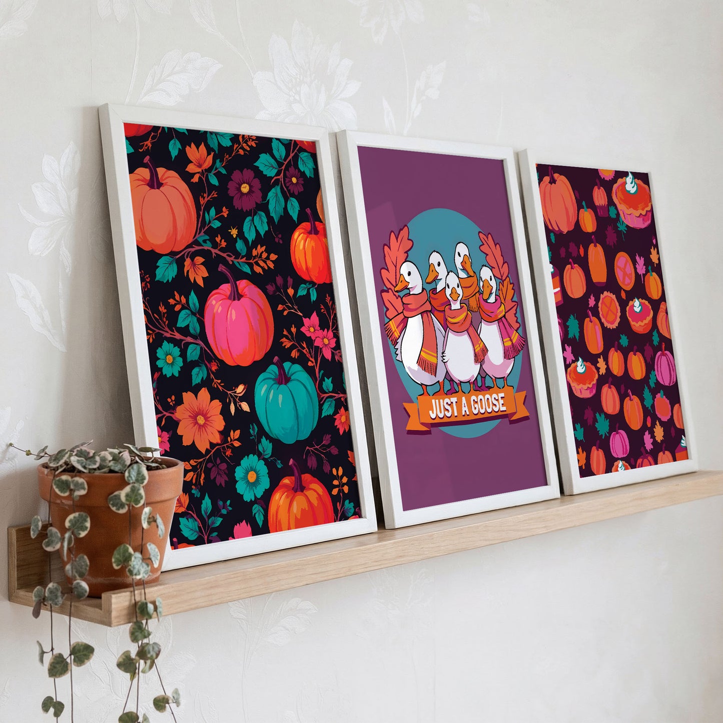 Styled shelf display of three maximalist autumn prints in white frames on a warm wood ledge - dark background pumpkin florals, scarfed geese on plum, and a deep berry-toned pumpkin pie pattern, with a trailing green plant