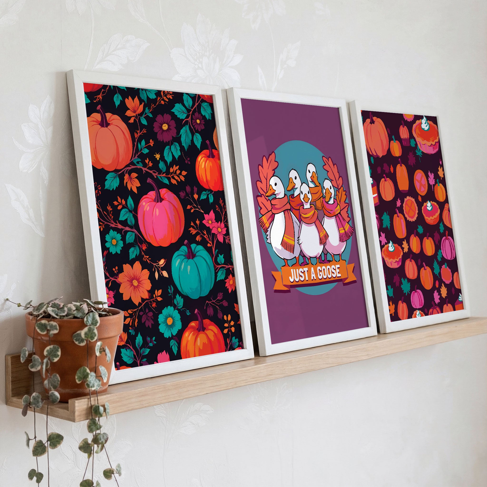 Styled shelf display of three maximalist autumn prints in white frames on a warm wood ledge - dark background pumpkin florals, scarfed geese on plum, and a deep berry-toned pumpkin pie pattern, with a trailing green plant