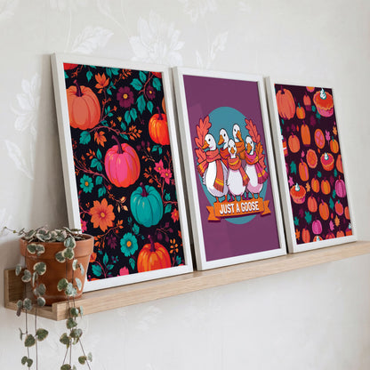 Styled shelf display of three maximalist autumn prints in white frames on a warm wood ledge - dark background pumpkin florals, scarfed geese on plum, and a deep berry-toned pumpkin pie pattern, with a trailing green plant