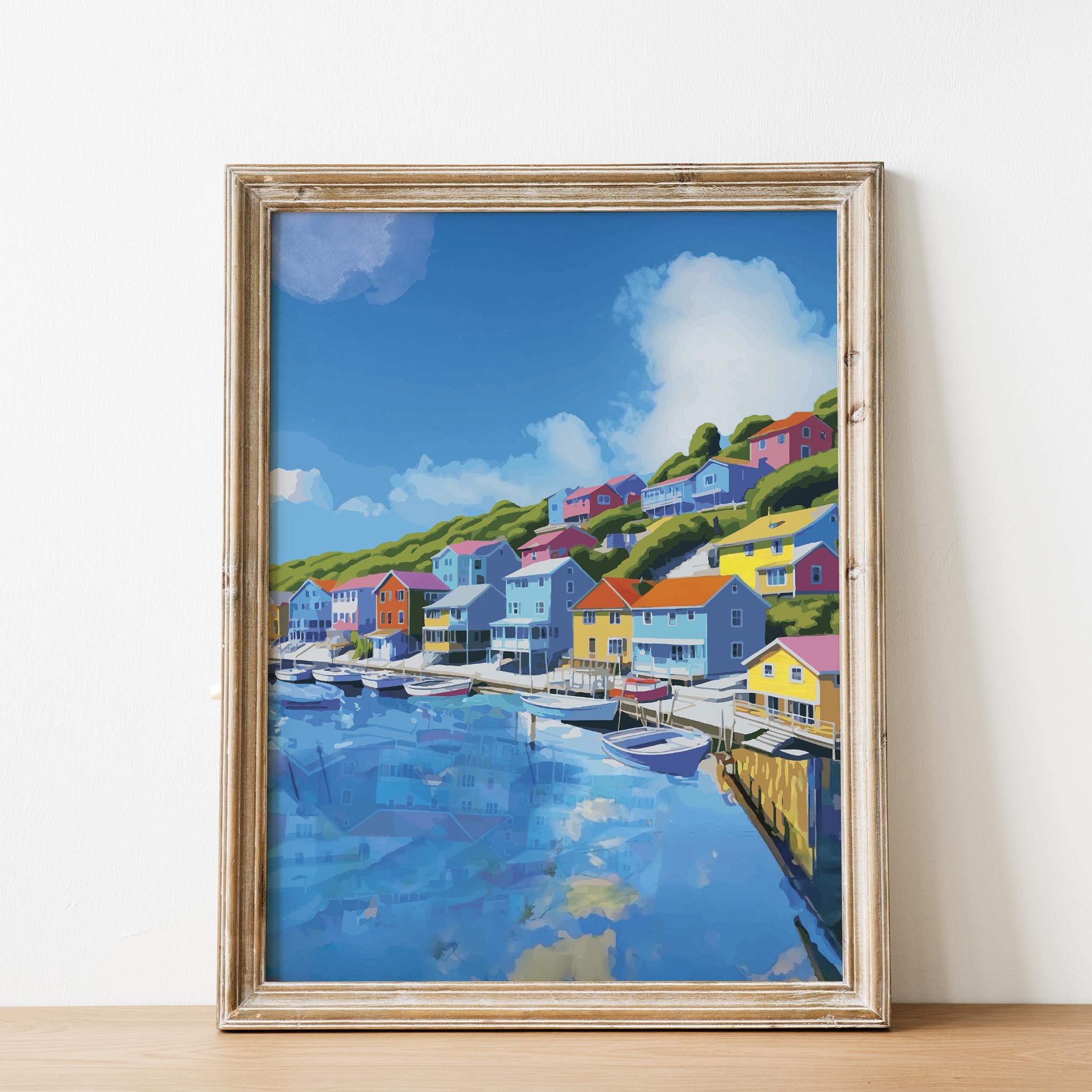 Charming harbor town wall art featuring vibrant seaside houses in bold colors lining a hillside above a marina filled with boats, under a bright blue sky with fluffy clouds reflected in the water