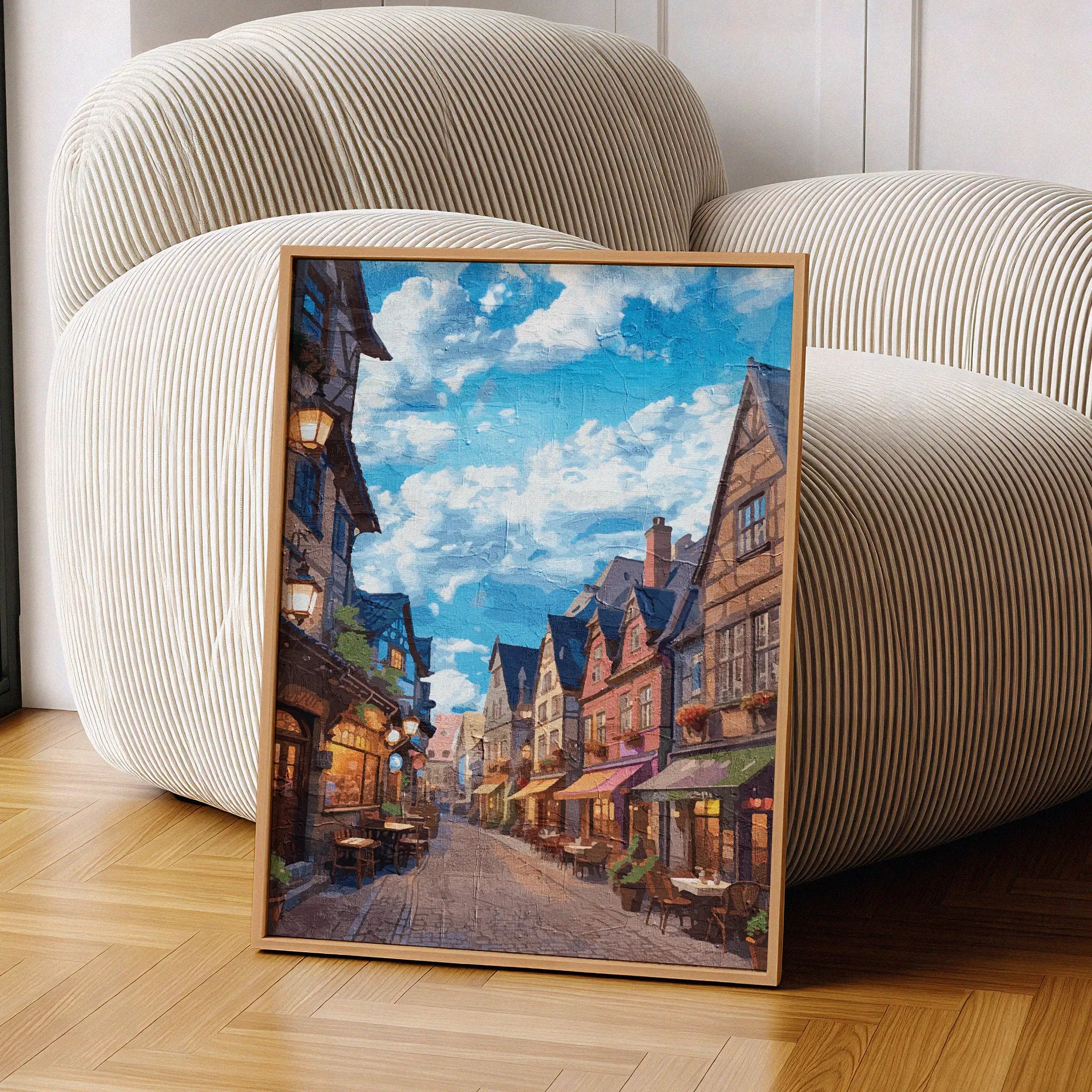 Cozy European village street wall art depicting a cobblestone alley lined with colorful historic buildings, quaint outdoor cafés, and warm lights under a vibrant blue sky with scattered clouds