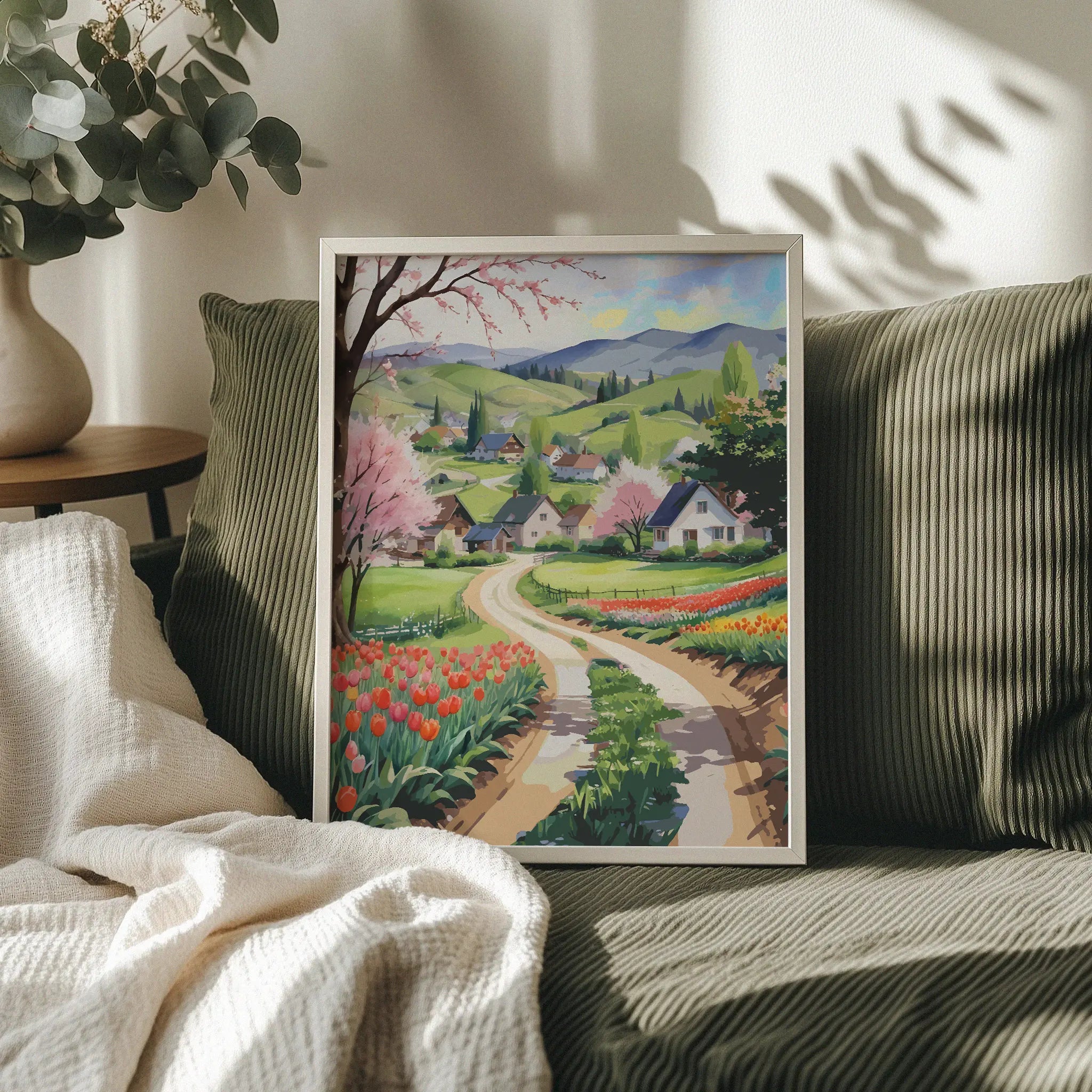 Charming countryside printable wall art showcasing a winding dirt road through vibrant tulip fields, blossoming trees, and cozy houses nestled in green rolling hills with distant mountains, perfect for spring, farmhouse, or cottagecore-inspired decor