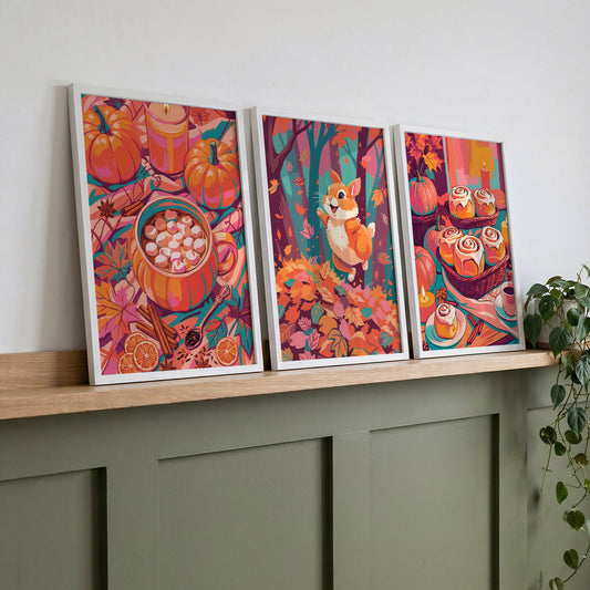 Three white-framed dopamine-style prints leaning on a natural oak ledge with a trailing plant, featuring cozy autumn scenes of spiced drinks, a joyful bunny, and iced cinnamon rolls