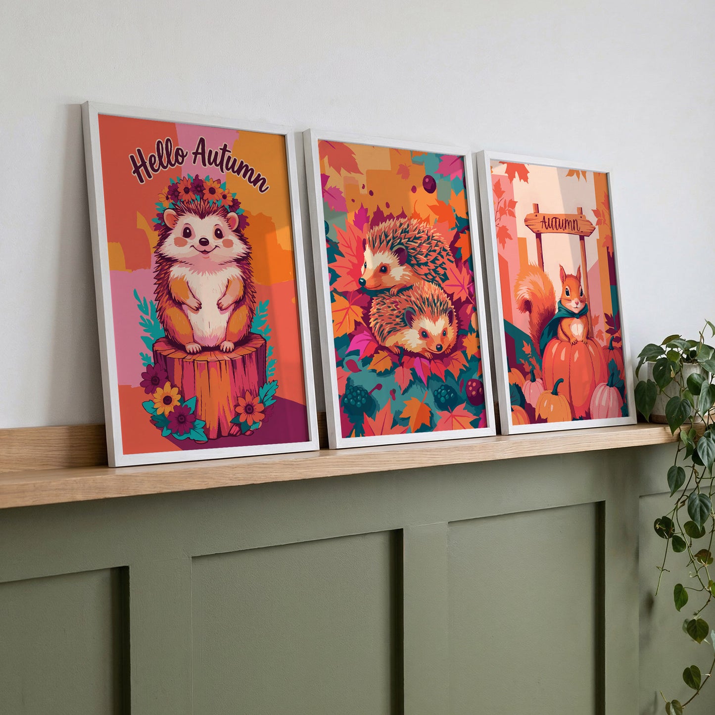 Three autumn woodland animal prints on a wood shelf against sage green panelling - a "Hello Autumn" hedgehog with flower crown, a hedgehog pair in fall leaves, and a teal-caped squirrel on a pumpkin patch