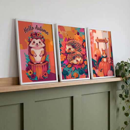 Three autumn woodland animal prints on a wood shelf against sage green panelling - a "Hello Autumn" hedgehog with flower crown, a hedgehog pair in fall leaves, and a teal-caped squirrel on a pumpkin patch