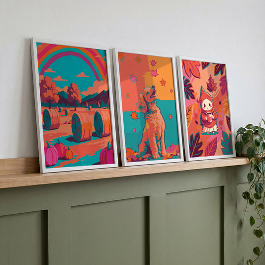 Three bold autumn wall art prints on a wood shelf against sage green panelling - a rainbow harvest landscape, a dog portrait, and a kawaii autumn gnome print in vibrant orange, teal, and pink