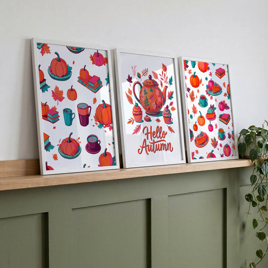 Three framed autumn wall art prints leaning on a wood shelf against a sage green panelled wall - featuring cozy fall pattern prints and a "Hello Autumn" teapot illustration in orange, pink, and teal
