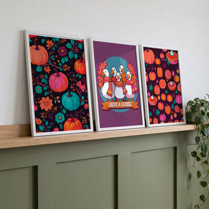 Three framed bold autumn wall art prints on a wood shelf against sage green panelling - a dark floral pumpkin pattern, a "Just a Goose" goose squad print, and a deep plum pumpkin pie pattern in saturated teal, orange, and magenta