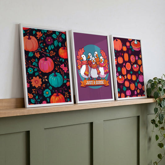 Three framed bold autumn wall art prints on a wood shelf against sage green panelling - a dark floral pumpkin pattern, a "Just a Goose" goose squad print, and a deep plum pumpkin pie pattern in saturated teal, orange, and magenta