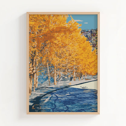 Autumn city park wall art featuring a row of golden-yellow trees along a riverbank, with reflections in the water and a hillside town in the distance under a clear blue sky